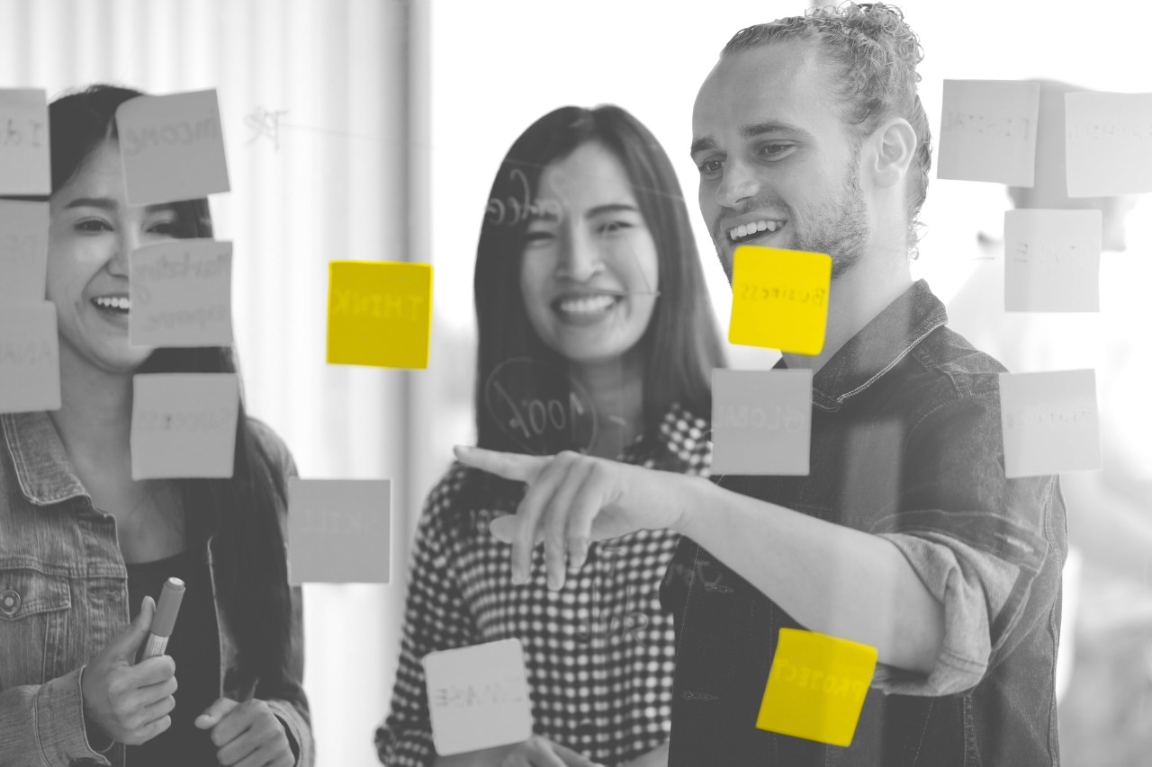 Group of young successful creative multiethnic team smile and brainstorm on project together in modern office with post note or sticky note. Caucasian man point on glass wall sharing idea. Group of young successful creative multiethnic team smile and brainstorm on project together in modern office with post note or sticky note. Caucasian man point on glass wall sharing idea.